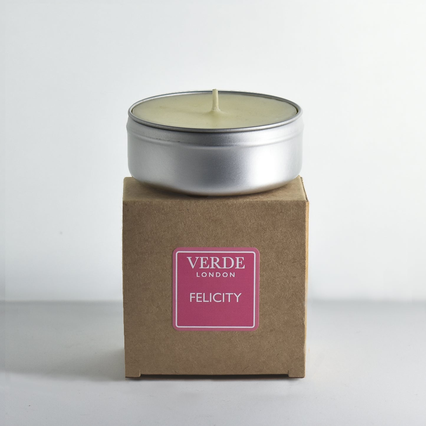 Candle in a metal tin on a cardboard box with 'Verde London Felicity' pink label on a white background