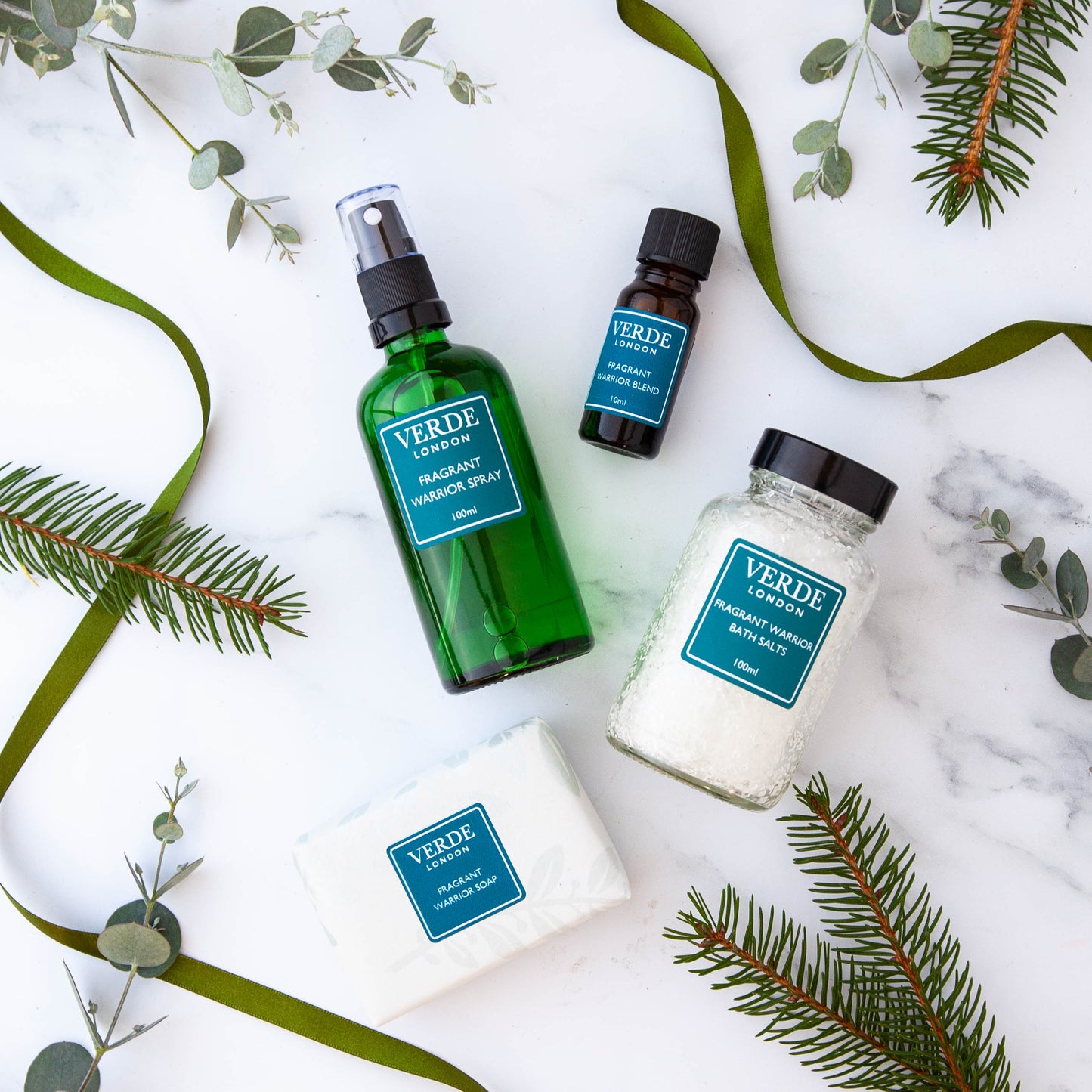 A collection of Fragrant Warrior VERDE branded products including a green spray bottle, a brown oil blend bottle, a white container of bath salts, and a soap, all arranged on a marble surface with decorative greenery in the background.