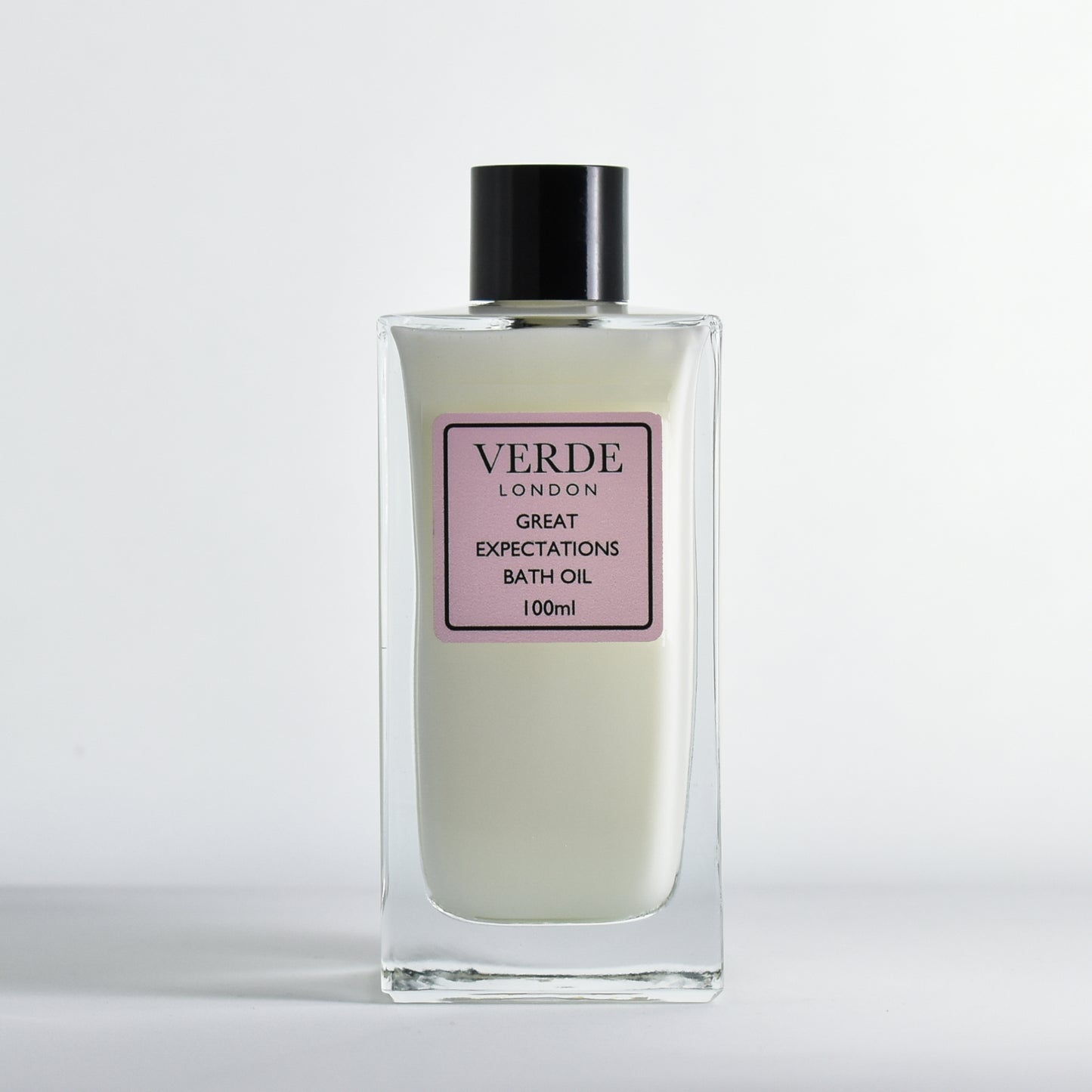 Bottle of Verde London 'Great Expectations' bath oil, a white milky colour with a pale pink label in a rectangular glass bottle on a white background