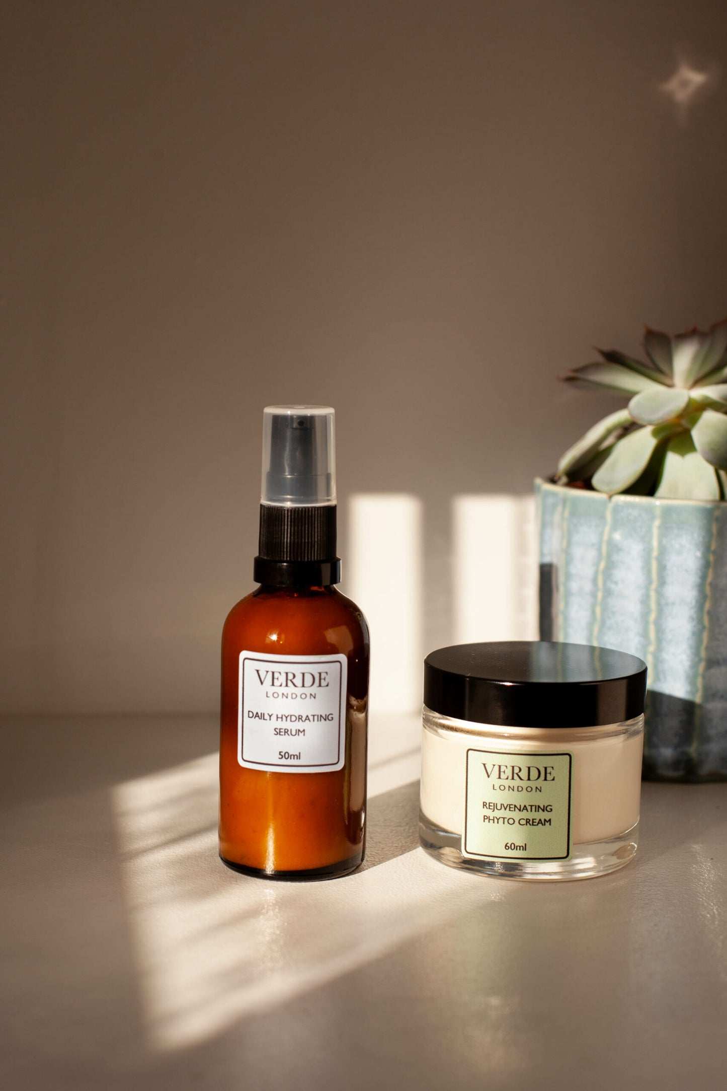 Bottle of daily hydrating serum and a glass jar of rejuvenating phytocream by the band Verde London on a shelf with a plant in the background