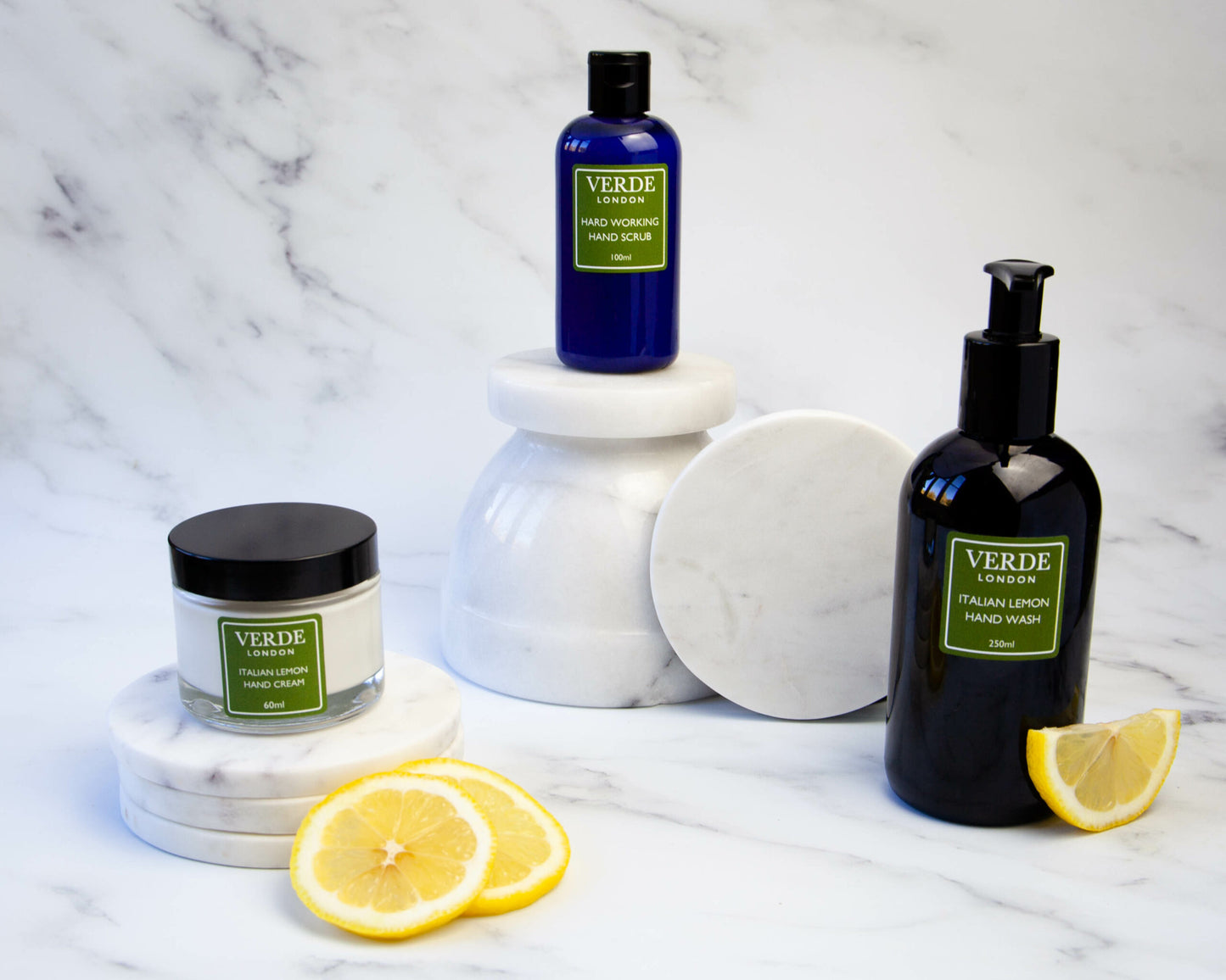 A collection of Verde London branded skincare products, including a bottle with a pump of hand wash, a glass jar of hand cream, and a bottle of hand scrub, displayed on a marble surface with lemon slices.