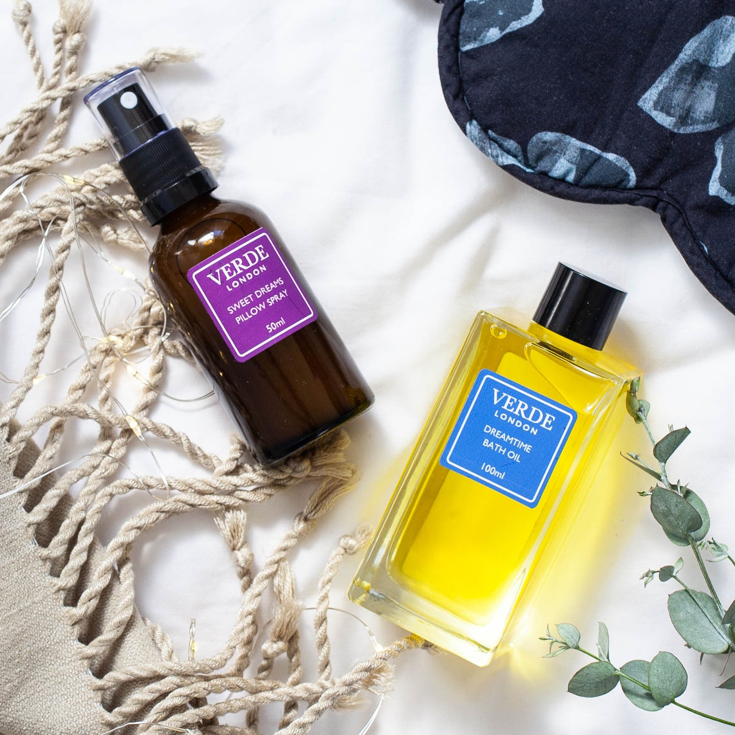 Two bottles of Verde London products, one Sweet dreams pillow spray in an amber glass spray bottle with a purple label and a clear glass rectangular bottle of Dreamtime Bath Oil with a blue label on a white sheet with eye mask a towel and some foliage