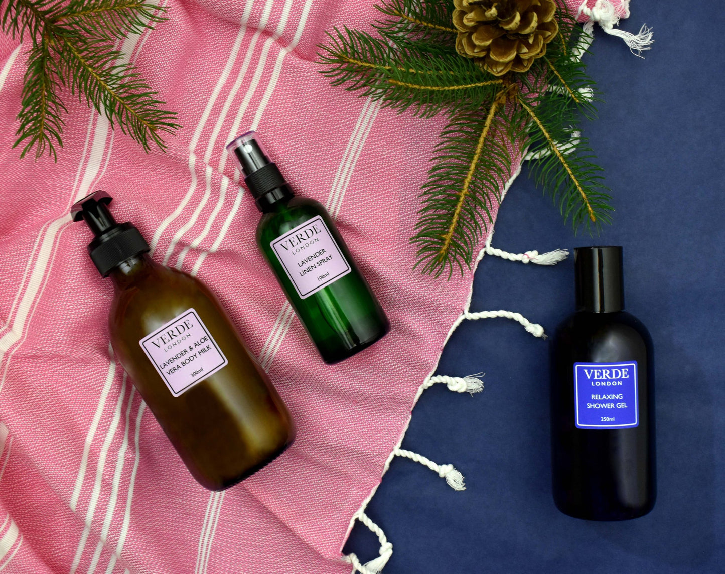 Three Verde branded bottles of bath and body products, including a shower gel, body milk, and linen spray, displayed on a textured fabric background with pine cones and a striped fabric at the top.