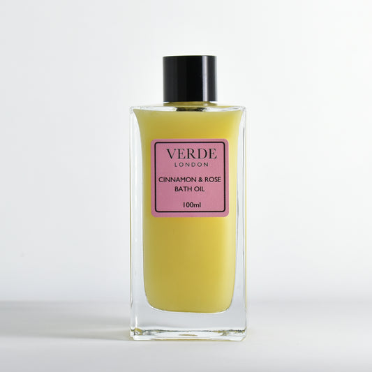 Bottle of 100ml Cinnamon and Rose Bath oil, milky yellow in colour with a pink label in a rectangular glass bottle on a white background