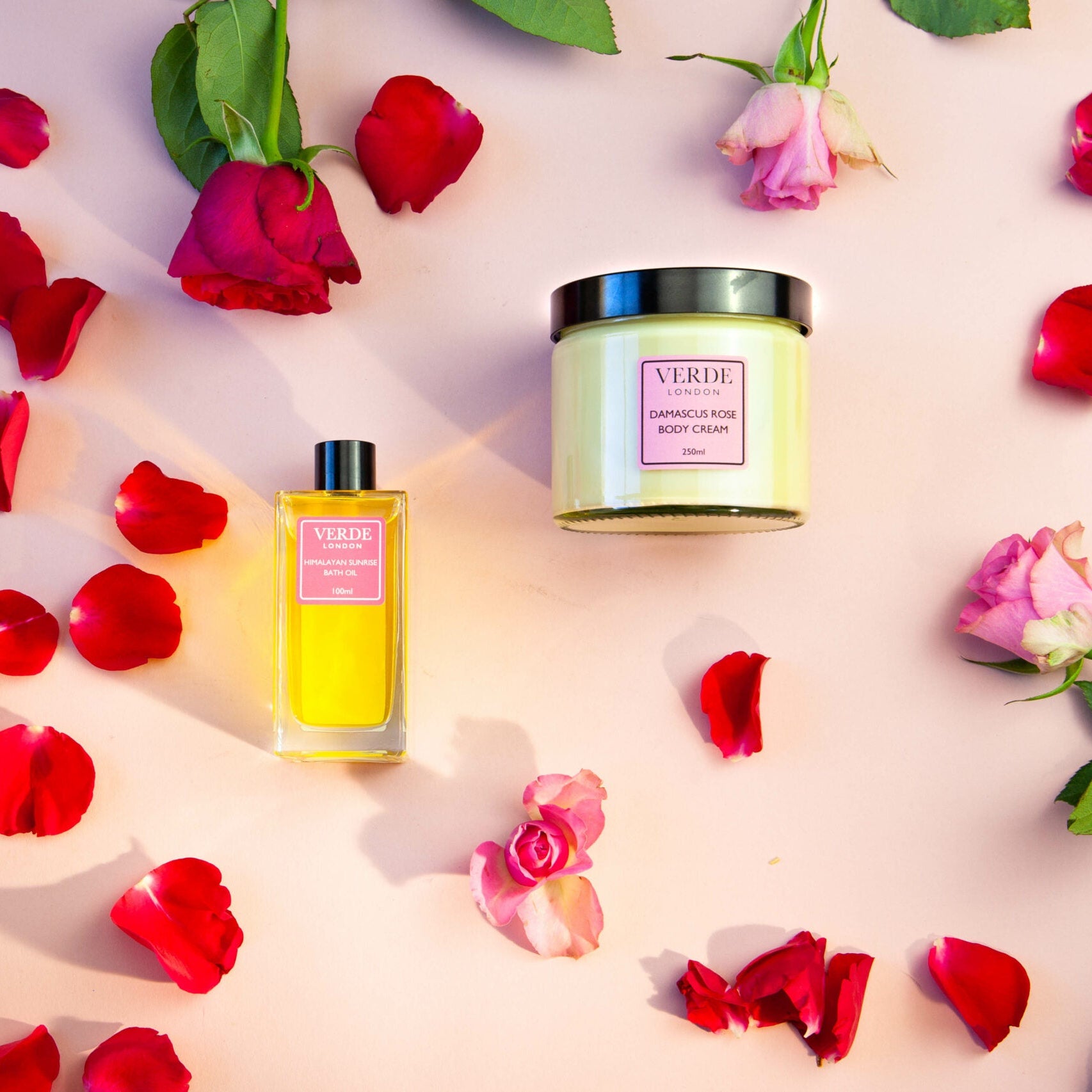 Two Verde London products on a pink background with rose petals. One is a bath oil in a clear rectangular bottle and the other is a clear glass jar both with pink labels on.