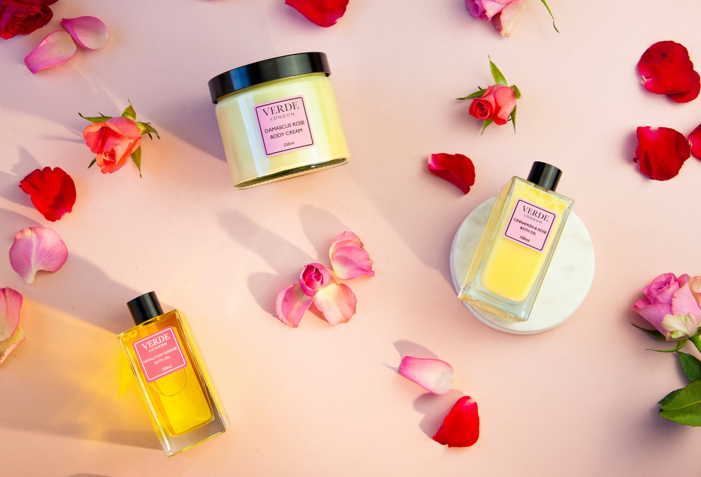 Three Verde London bath and body products including two bath oils in rectangular glass bottles and one large jar of body cream all on a pink background with rose petals 