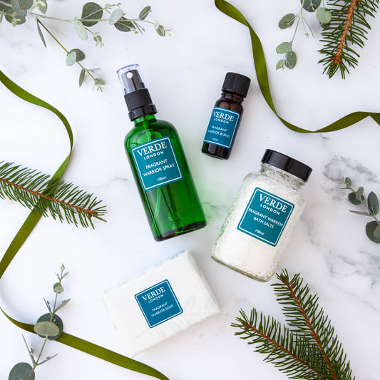 A collection of Fragrant Warrior VERDE branded products including a green spray bottle, a brown oil blend bottle, a white container of bath salts, and a soap, all arranged on a marble surface with decorative greenery in the background.