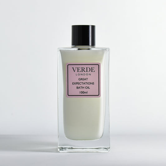 Bottle of Verde London 'Great Expectations' bath oil, a white milky colour with a pale pink label in a rectangular glass bottle on a white background