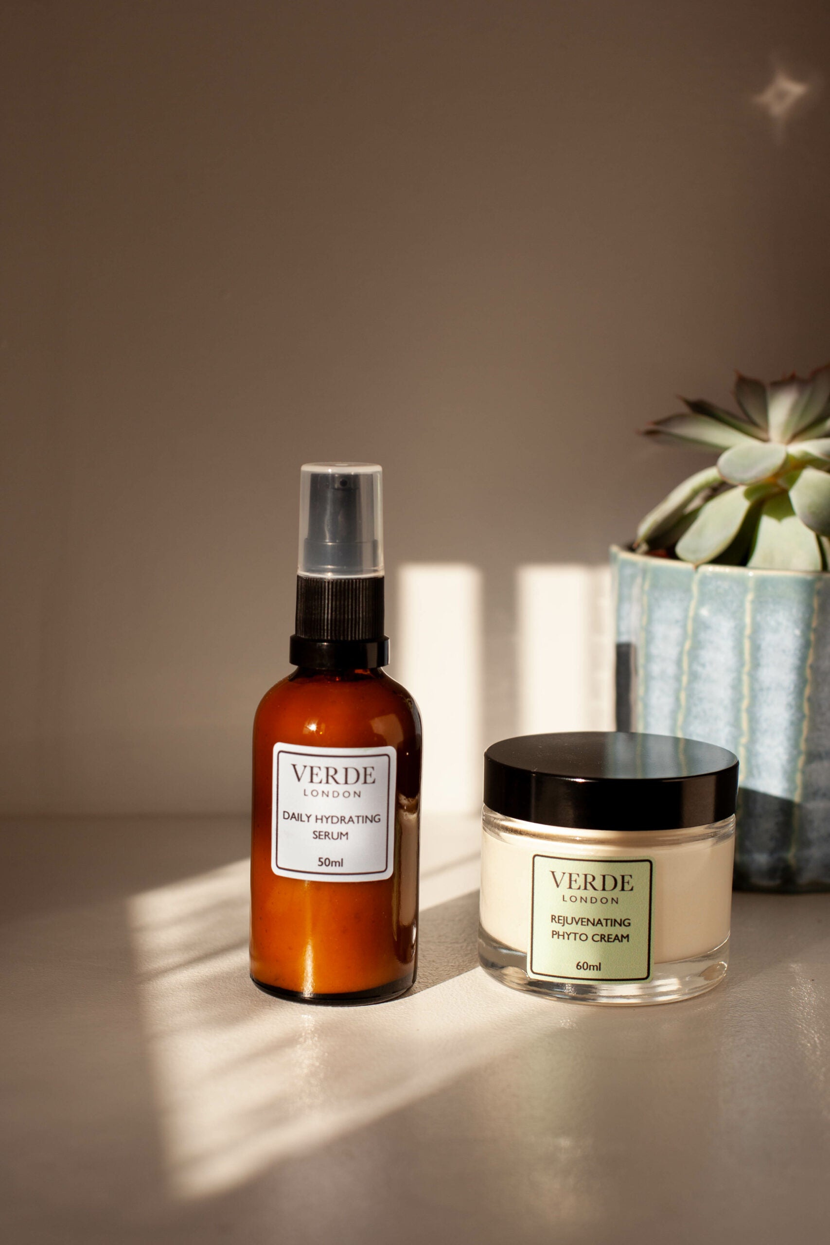 Bottle of daily hydrating serum and a glass jar of rejuvenating phytocream by the band Verde London on a shelf with a plant in the background
