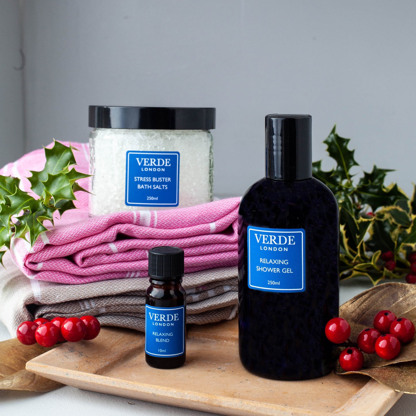 A gift pack from VERDE including a jar of bath salts, a pump bottle of shower gel, and a small bottle of oil, all displayed on a wooden tray with holly and berries in the background.