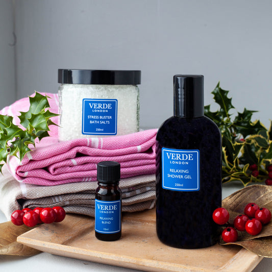 A gift pack from VERDE including a jar of bath salts, a pump bottle of shower gel, and a small bottle of oil, all displayed on a wooden tray with holly and berries in the background.