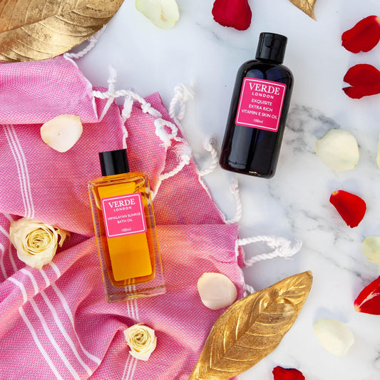 A bottle of Exquisite Extra Rich Vitamin E Body Oil and a glass bottle of Himalayan Sunrise Bath Oil both by the brand Verde London with pink labels on a marble background with a pink towel and rose petals.