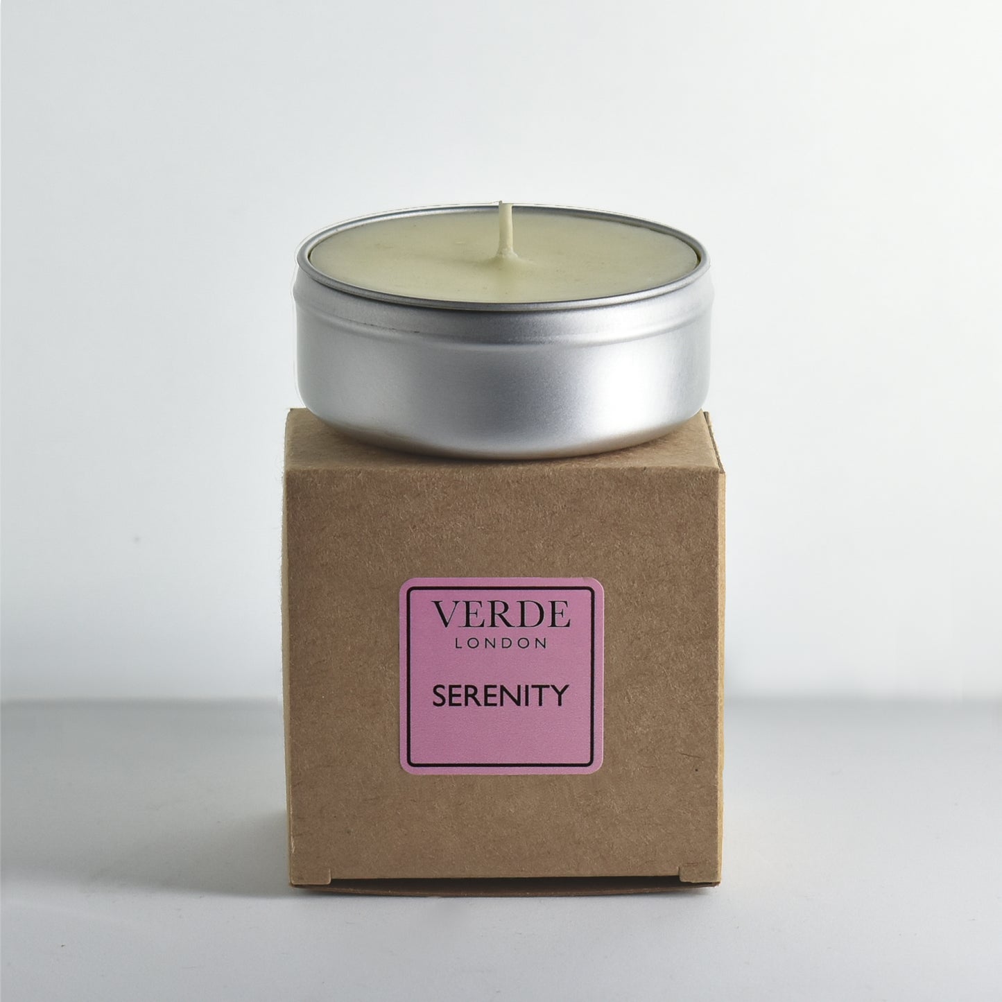 Candle in a metal tin on a cardboard box with 'Verde London Serenity' pink label.