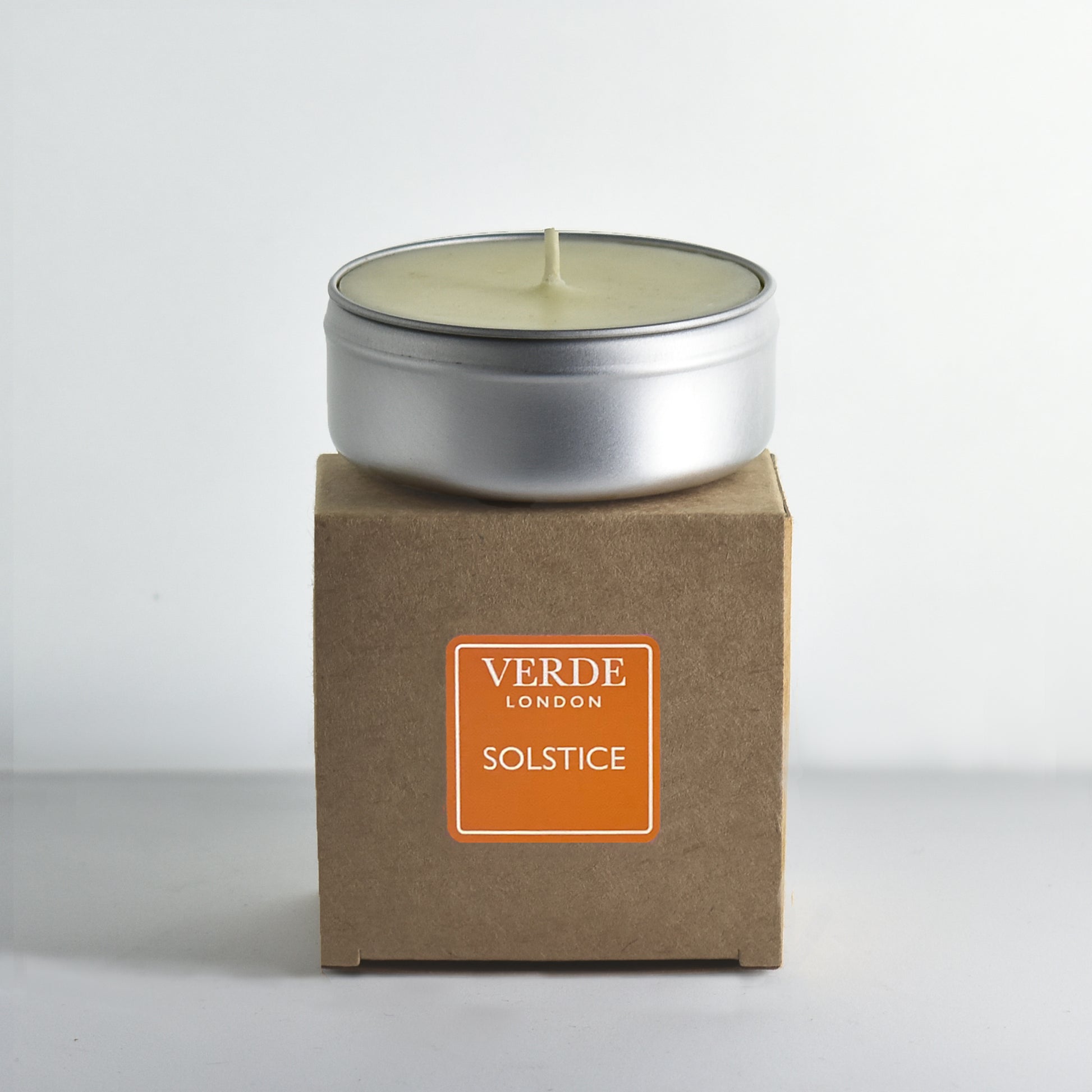 Candle in a metal tin on a cardboard box with and orange 'Verde London Solstice' label on a white background