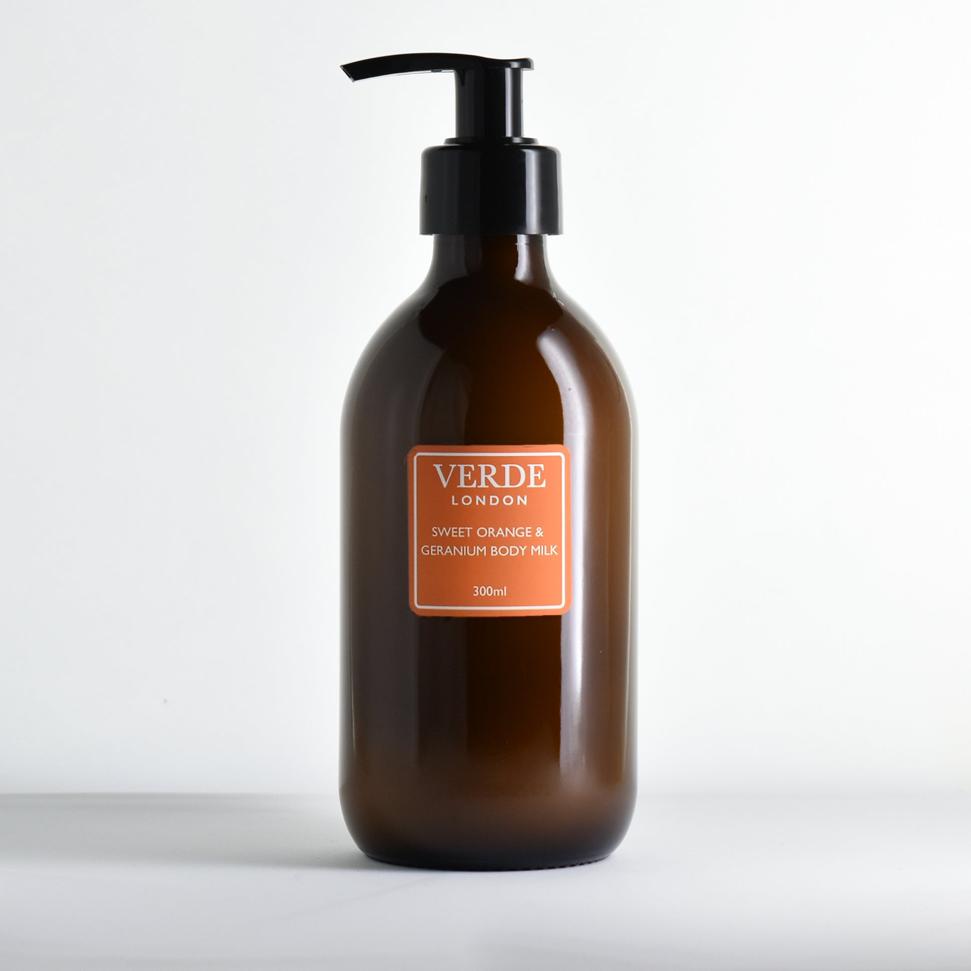Amber bottle with pump of Sweet Orange and Geranium Body Milk with an orange label by the brand 'Verde London' on a white background