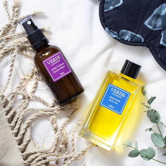 Two bottles of Verde London products, one Sweet dreams pillow spray in an amber glass spray bottle with a purple label and a clear glass rectangular bottle of Dreamtime Bath Oil with a blue label on a white sheet with eye mask a towel and some foliage
