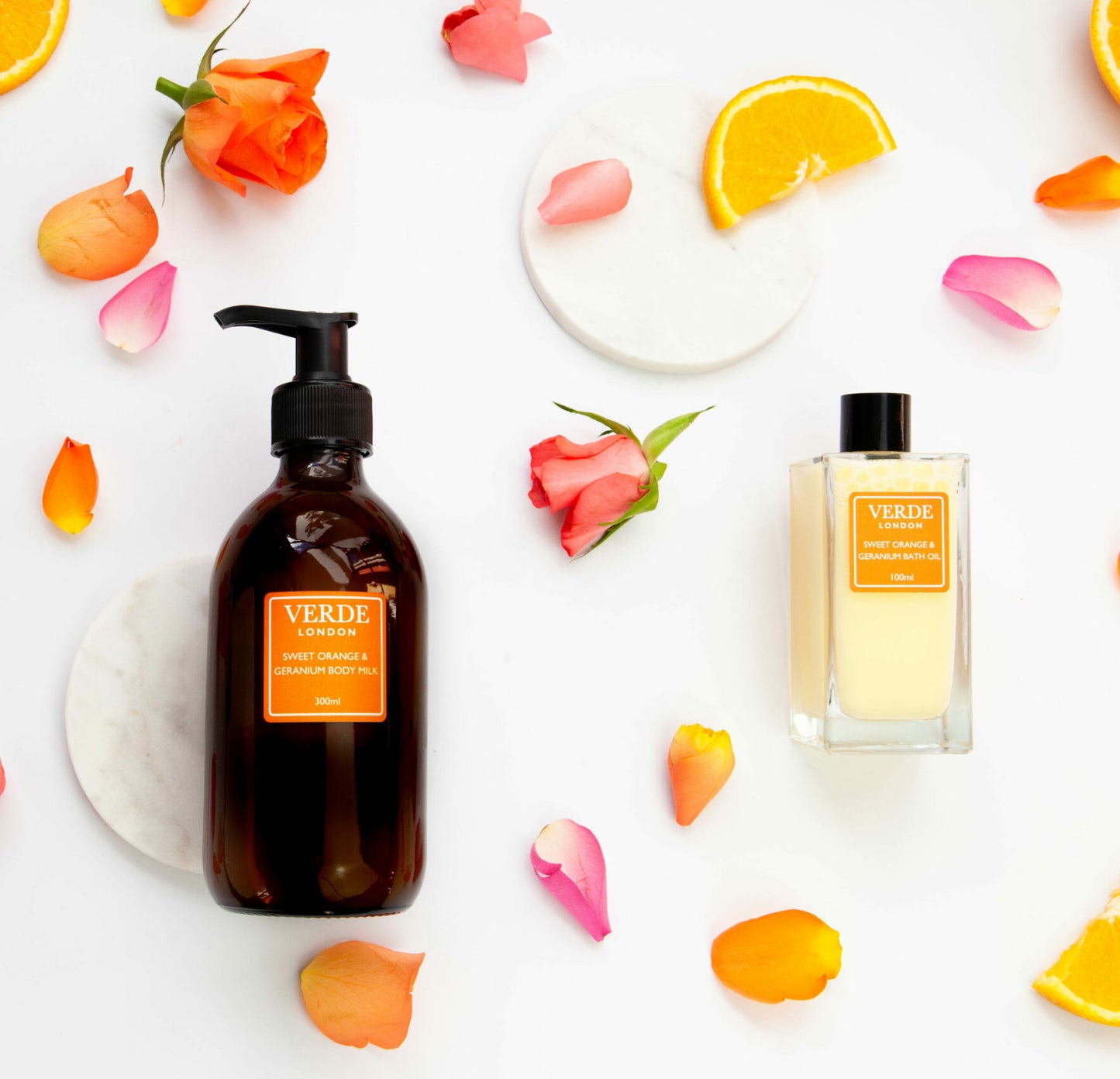 Amber glass pump bottle and clear rectangular bottle of Sweet Orange and Geranium body milk and bath oil with orange labels on a white background with rose petals and orange slices
