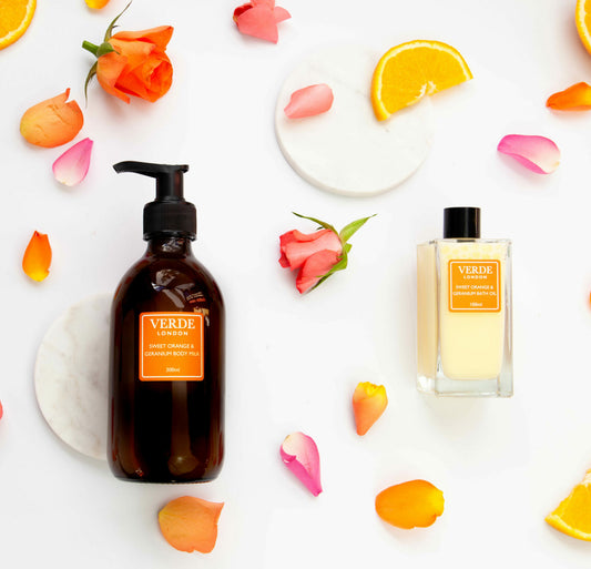 Amber glass pump bottle and clear rectangular bottle of Sweet Orange and Geranium body milk and bath oil with orange labels on a white background with rose petals and orange slices