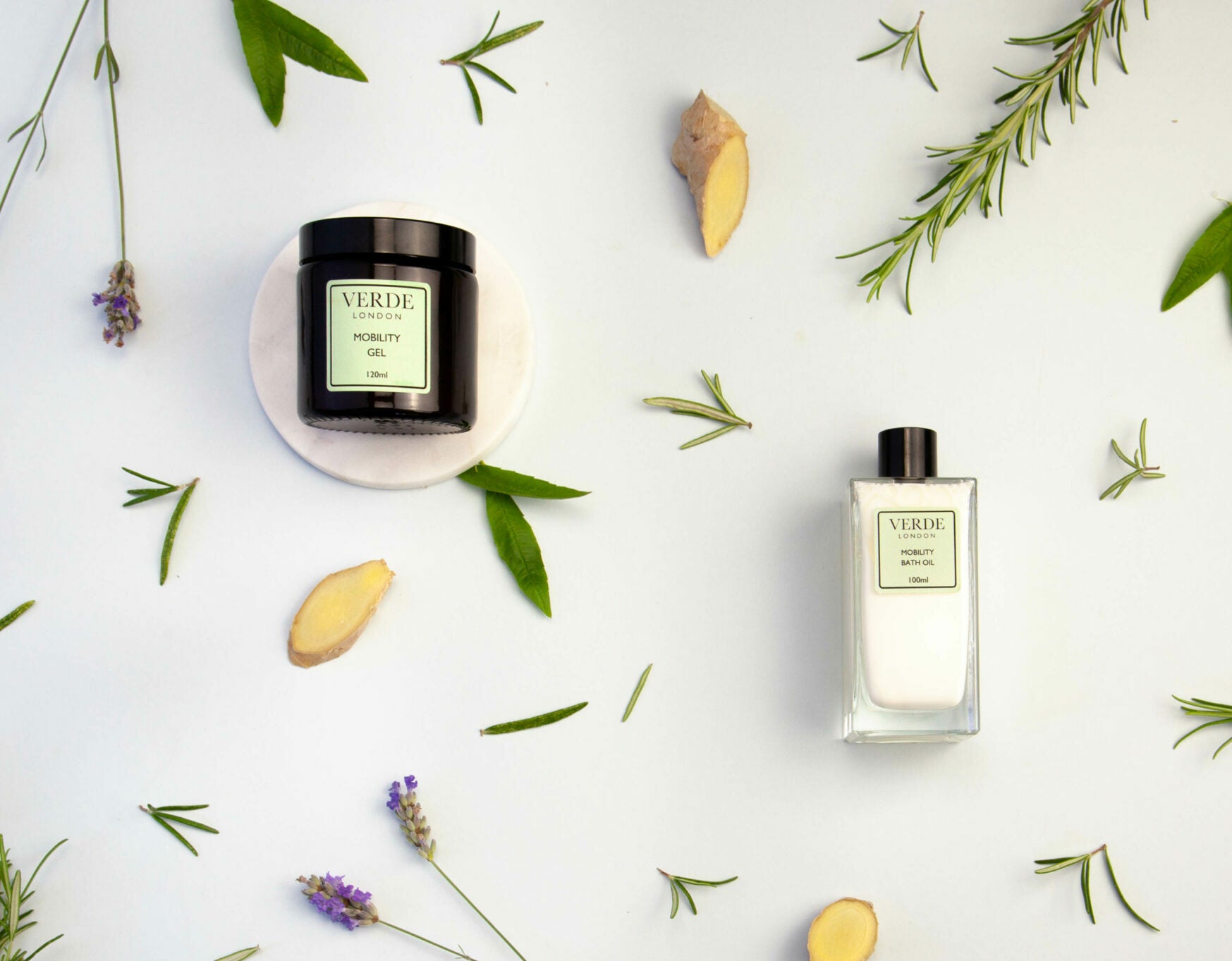 Two bottles of Verde London branded products, a Mobility Gel and a Mobility Bath Oil, one with in an amber glass jar and the other in a rectangular glass bottle, surrounded by decorative greenery and a few dried plant elements on a white background.