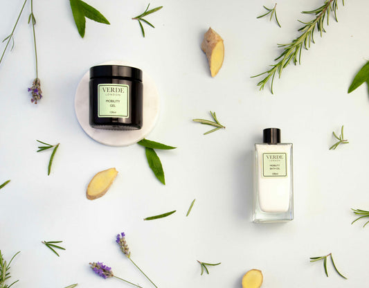 Two bottles of Verde London branded products, a Mobility Gel and a Mobility Bath Oil, one with in an amber glass jar and the other in a rectangular glass bottle, surrounded by decorative greenery and a few dried plant elements on a white background.
