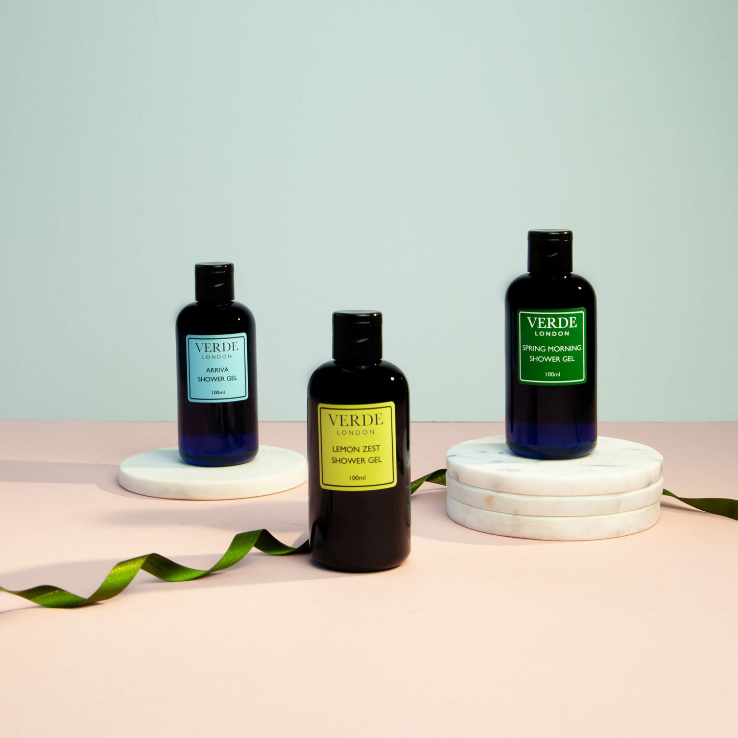 Three bottles of shower gel on white round bases with a label reading 'VERDE' and different scents indicated such as 'Arriva', 'Spring Morning', and 'Lemon Zest'.