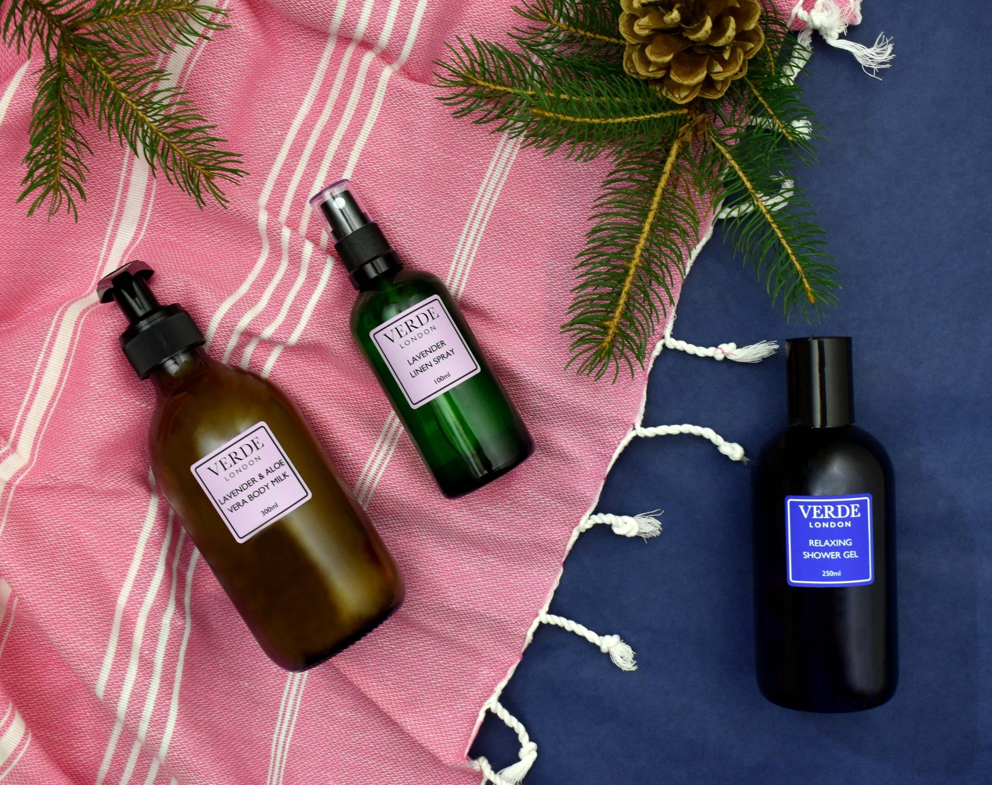Three Verde branded bottles of bath and body products, including a shower gel, body milk, and linen spray, displayed on a textured fabric background with pine cones and a striped fabric at the top.